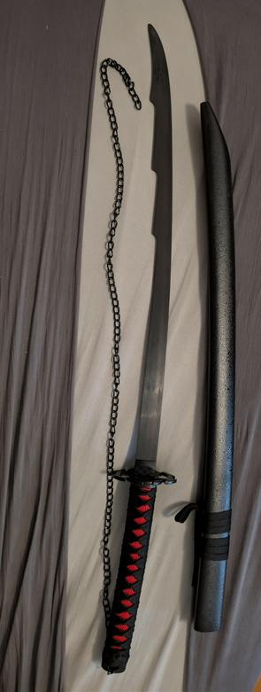 Vertikales Foto eines nachgebildeten Zanpakutō „Zangetsu“ in Ichigo Kurosakis Bankai-Form (auch „Tensa Zangetsu“) aus Bleach. Das Schwert liegt ungeschwärzt auf einem Bettbezug in Grau- und Beigetönen, die Spitze zeigt nach oben. Die Klinge ist schlank, leicht gebogen und in dunklem, matt schimmerndem Stahl gehalten; an der stumpfen Rückenlinie befinden sich dekorative Ausnehmungen nahe der Spitze, die dem Anime-Design nachempfunden sind. Am Griffende ist eine schwarze Metallkette befestigt, die locker S-förmig neben der Klinge verläuft. Der Griff (Tsuka) ist traditionell gewickelt: schwarze Umwicklung mit roten, rautenförmig sichtbaren Zwischenlagen. Die schwarze Parierstange (Tsuba) wirkt rund und minimalistisch, passend zur Bankai-Ästhetik. Rechts neben dem Schwert liegt die dazugehörige, ebenfalls schwarze Scheide (Saya) mit fein gesprenkelter Oberfläche und zwei Bändern zum Befestigen. Keine Personen im Bild, neutraler Hintergrund; Fokus liegt auf Material, Proportionen und charakteristischen Designelementen der Anime-Replik.