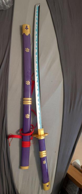 Auf einer grauen Bettdecke liegt eine günstige Holz-Replik des Königsschwerts „Enma“ von Lorenor Zorro aus One Piece. Das Katana ist vollständig aus der Scheide gezogen; Klinge und Scheide liegen parallel und direkt nebeneinander, quer durchs Bild von links (Spitze) nach rechts (Griff). Die Holz­klinge ist silbern bemalt, hat einen schwarzen Rücken und mittig eine türkisfarbene Bahn mit weißer, wellenförmiger Zierlinie im Stil einer Hamon. Der Griff (Tsuka) ist glatt violett lackiert, mit goldfarbenen Beschlägen; die runde, goldene Parierstange (Tsuba) und die goldene Zwinge wirken deutlich als Kunststoff/Spielzeug.

Die Schwertscheide (Saya) unterhalb der Klinge ist ebenfalls violett, glänzend lackiert und mit goldenen Details verziert: mehrere kleine, stilisierte Blütenembleme, zwei goldene Ringe nahe der Öffnung sowie eine goldene Endkappe am unteren Ende, in die ein violettes Kreuzmotiv eingelassen ist. Am Mund der Scheide ist ein leuchtend rotes Band befestigt und locker umgelegt. Die handwerkliche Ausführung und das Material (lackiertes Holz, einfache Bemalung) zeigen, dass es sich um ein günstiges Deko-/Cosplay-Exemplar handelt, nicht um eine echte Metallwaffe. Am rechten Bildrand sind Teile eines Holz­möbels sichtbar; sonst füllt die Bettdecke den Hintergrund ohne weitere Gegenstände.