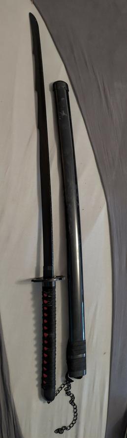 Auf einem grauweißen Stoffuntergrund liegen Scheide und Schwert des Zanpakutō Zangetsu in Ichigo Kurosakis Bankai-Form (Bleach) waagerecht nebeneinander. Es handelt sich um eine günstige Holzreplik: Die lange, komplett schwarz lackierte Klinge ist stumpf und leicht gebogen; nahe dem Griff ist ein kurzer Absatz angedeutet. Die Parierstange (Tsuba) ist schlicht und dunkel. Der Griff ist mit schwarzem Band in traditioneller Rautenwicklung umlegt, darunter schimmert eine rote Unterlage durch. Oberhalb liegt die glänzend schwarze Scheide (Saya) mit sichtbaren Gebrauchsspuren; am vorderen Ende ist sie mit Band umwickelt, daran hängt eine kurze Zierkette. Das minimalistische, durchgehend schwarze Design entspricht der Bankai-Ästhetik von Tensa Zangetsu; das Stück wirkt deutlich als Deko-/Cosplay-Variante ohne echte Schneide.