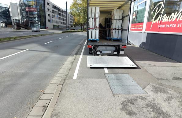 Ein LKW mit heruntergelassener Ladeklappe steht auf dem Bürgersteig neben einer Straße mit 2 Fahrstreifen pro Richtung. Auf dem Bürgersteig ist ein Radweg aufgemalt, den der LKW vollständig blockiert.
