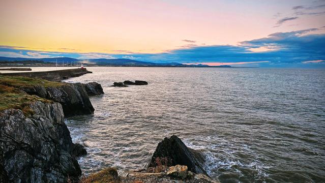 A dramatic coastal landscape at sunset. In the foreground on the left are dark, rugged cliffs dropping sharply to the turbulent sea. The middle ground is dominated by the dark, choppy water of the ocean stretching to the horizon. In the distance to the left, a small harbour wall with a white and red lighthouse is visible. The background features rolling hills under a vibrant sky with pastel pink, orange, and soft blue cloud cover. The overall mood is rugged yet serene.