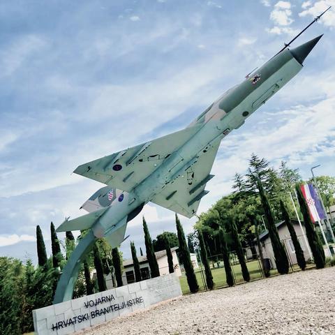 Croatian Air Force MiG-21 fighter jet monument mounted vertically on a stone base with text "VOJARNA HRVATSKI BRANITELJI ISTRE," surrounded by cypress trees.