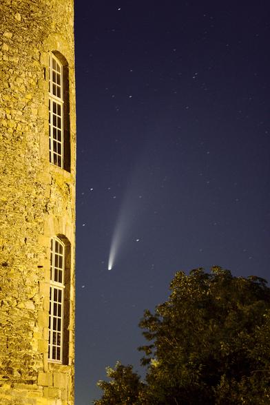 Photo en format vertical de la comète Neowise dans le ciel bleu nuit parsemé d'étoiles, avec une tour de château au premier plan à gauche de l'image. La comète semble plonger vers une fenêtre de la tour dont le mur est jaune vif, coloré par l'éclairage public.
