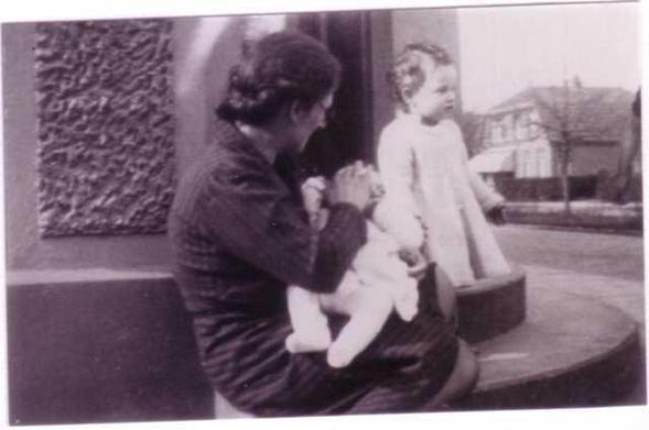 A mother sirring outside with her two children. A younger boy is in her arms, a little older girl is standing next to her.