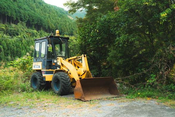 A yellow backhoe loader parked on a grassy patch, surrounded by dense green foliage and trees. The vehicle displays the brand label "Yanmar V4-6". A forested hillside forms the background under a cloudy sky.