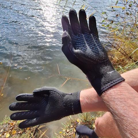 Zwei Hände mit Neopren-Handschuhen. Darunter ein Fuß mit Neopren-Socke. Noch auf dem Seeufer stehend. In Hintergrund eine Wasseroberfläche, die von der Sonne beschienen wird.