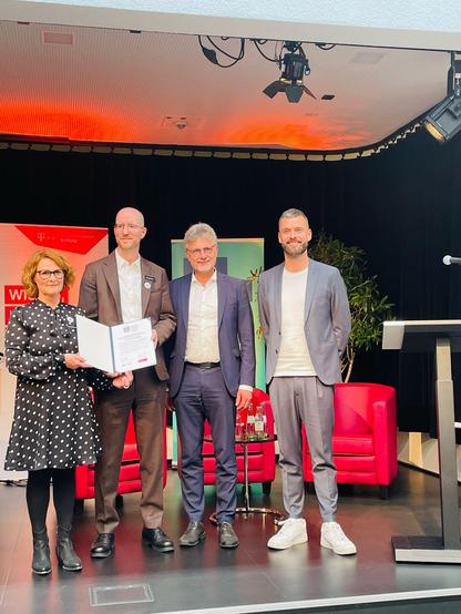 Drei Männer und eine Frau stehen auf der Bühne. Die Frau hält eine Urkunde.