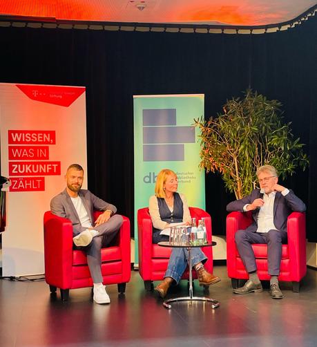 Drei Personen sitzen in roten Sesseln auf der Bühne.