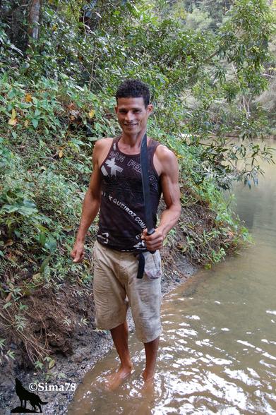 Homme souriant tenant une machette, debout dans une rivière entourée de végétation tropicale.
Smiling man holding a machete, standing in a river surrounded by tropical vegetation.
Hombre sonriente con un machete en la mano, de pie en un río rodeado de vegetación tropical.
