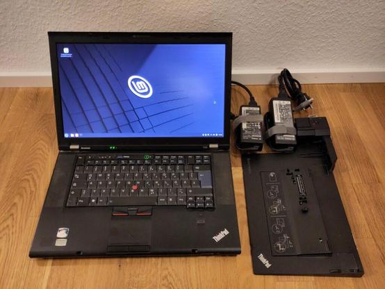 Foto des laufenden Laptops mit üblichen Gebrauchsspuren, das den Linux Mint-Desktop anzeigt. Daneben liegen ein Dock und 2 Netzteile.