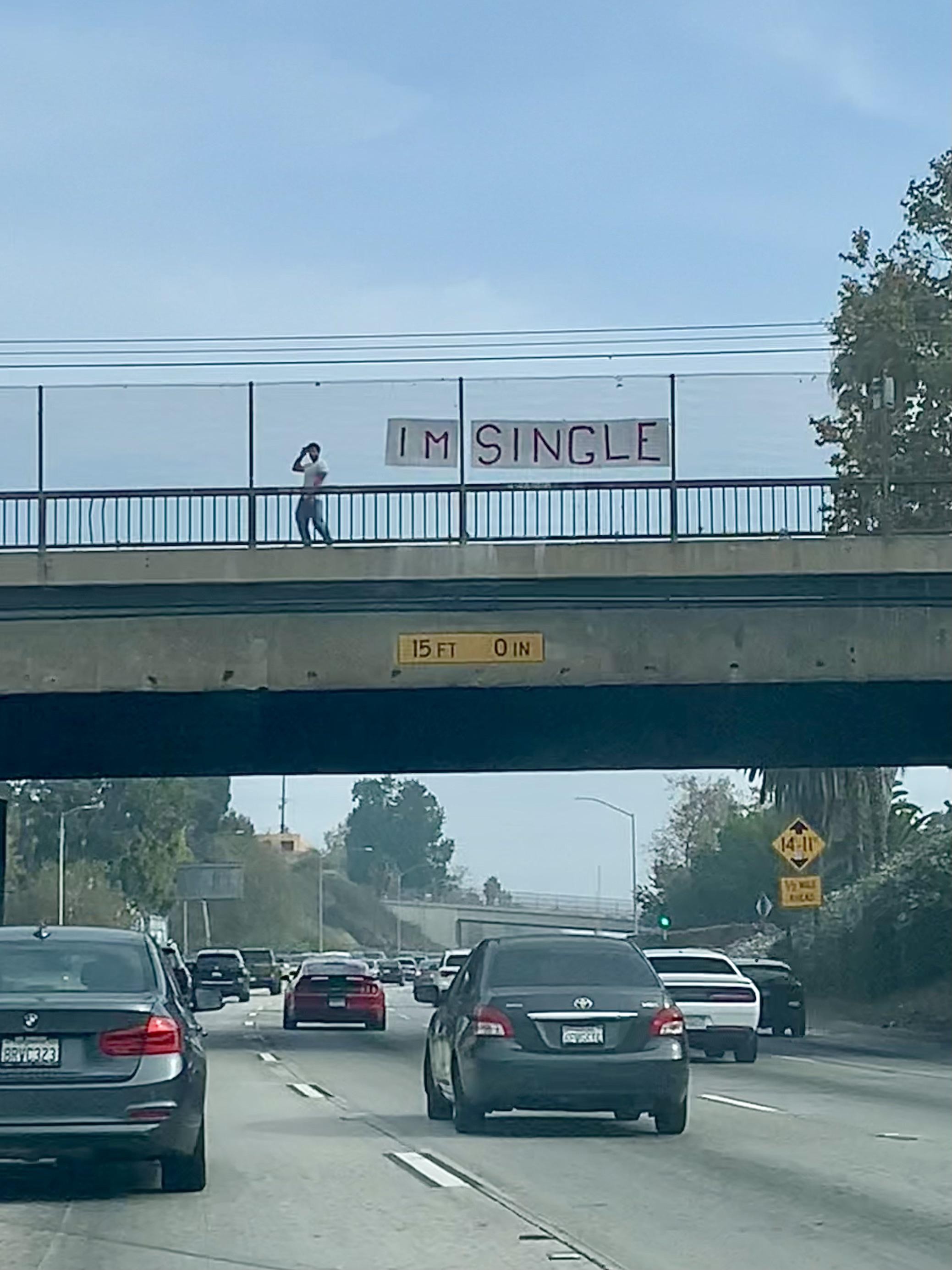 A photo of a freeway overpass.  The traffic is moderate, the sky is blue.  A man in white t-shirt and jeans strikes a pose next to a large banner with "I'm Single" in hand drawn red block lettering.