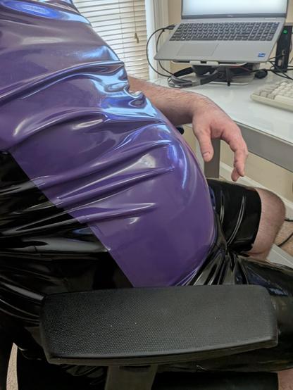 Photo of me sitting in my work from home chair with my black latex and purple t-shirt on and black latex shorts