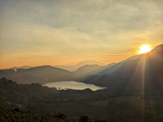 This image captures a breathtaking landscape at sunrise or sunset. The sun is low in the sky, casting a warm, golden glow across the scene. Rolling hills and mountains stretch into the distance, their silhouettes softened by the gentle light. A serene lake sits nestled between the hills, reflecting the soft hues of the sky. The foreground features a mix of lush greenery and open fields, adding depth and texture to the scene. The sun's rays create a striking lens flare effect, adding a magical touch to the tranquil atmosphere. The overall mood is peaceful and calming, evoking a sense of serenity and natural beauty.