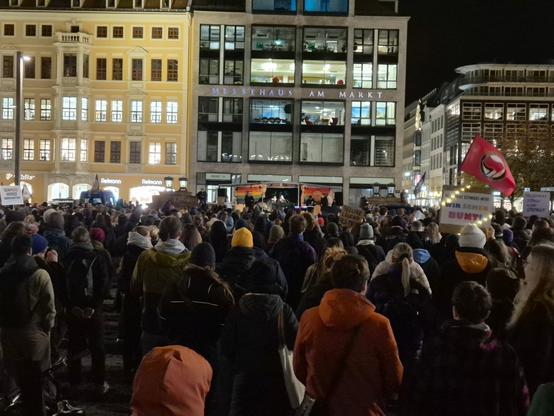 Eine Menschenmenge versammelt sich auf einem öffentlichen Platz in einer Stadt bei Nacht. Die Beleuchtung ist hell, vor allem von den umliegenden Gebäuden. Im Hintergrund dominieren zwei große Gebäude: links ein hellgelb gestrichenes, traditionell aussehendes Gebäude mit mehreren Reihen von Fenstern und rechts ein modernes, größeres Bürogebäude mit großen rechteckigen Fenstern, das prominent die Aufschrift "MESSEHAUS AM MARKT" trägt. Das Messehaus ist ebenfalls gut beleuchtet und weist mehrere beleuchtete Büroräume auf.
Der Platz ist gefüllt mit einer großen Ansammlung von Menschen, die alle zum Zentrum des Platzes blicken, wo sich eine kleine Gruppe auf einem Podest oder einer temporären Bühne zu versammeln scheint. Die Versammlung blickt von hinten auf die Beobachter:innen, was einen Eindruck der Tiefe der Menge vermittelt. Einige Personen in der Menge tragen auffällige Kleidung wie eine gelbe Strickmütze oder eine leuchtend orangefarbene Kapuzenjacke.
Auf der rechten Seite der Menge ist eine Person zu erkennen, die eine rote Fahne mit einem schwarzen Emblem hält. Mehrere Schilder oder Plakate sind in der Menge sichtbar, darunter eines rechts, das auf Deutsch "WIR SEHEN DEINE FARBEN, DU SIEHST UNS BUNT!" zu lauten scheint, und ein anderes ganz links, auf dem nur teilweise "SOZIALE GRENZEN ÜBERWINDEN" zu erkennen ist. Die Stimmung scheint die einer organisierten, öffentlichen Demonstration oder Kundgebung zu sein. Die Nacht ist klar und die Stadtbeleuchtung ist dominant.

En