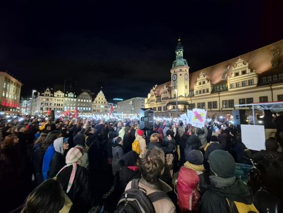 Ein nächtliches Foto zeigt eine große Menschenmenge, die sich auf einem Platz vor dem Alten Rathaus in Leipzig versammelt hat, im Rahmen einer Demonstration. Die Szene ist bei Dunkelheit aufgenommen, und das Rathaus sowie einige umliegende Gebäude sind beleuchtet. Das Alte Rathaus im Hintergrund ist ein hellgelb gestrichenes Gebäude mit einem charakteristischen Turm in der Mitte, der eine Uhr und ein grünes Dach hat. Rechts vom Turm ist das Gebäude braun-rot gehalten. Die Menschenmenge füllt den vorderen und mittleren Bereich des Bildes vollständig aus. Viele Personen in der Menge halten ihre Mobiltelefone hoch, wodurch zahlreiche helle Punkte in der dunklen Szene entstehen. Einige Menschen tragen Mützen, Schals und dicke Jacken in verschiedenen Farben, was auf kühles Wetter hinweist. Im Zentrum der Menge steht eine dunkle Lautsprecheranlage auf einem Podest. Einzelne Schilder und Plakate sind in der Menge zu erkennen, zum Beispiel ein Plakat mit einer deutschen Flagge und einem stilisierten Herz. Im rechten Bildvordergrund sind hauptsächlich die Hinterköpfe und Schultern der Teilnehmer:innen zu sehen, darunter eine Person mit einem beigefarbenen Rucksack und eine andere mit einem roten Rucksack.

Englische Zusammenfassung: A nighttime photo shows a large crowd of people gathered in front of the Old Town Hall in Leipzig, likely at a demonstration. The Old Town Hall with its characteristic tower is brightly illuminated in the background. The crowd fills the foreground and midd
