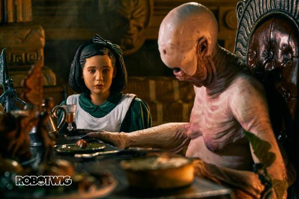 Ofelia approaches the Pale Man who sits on a big chair at a table full of food with its eyes on a plate.