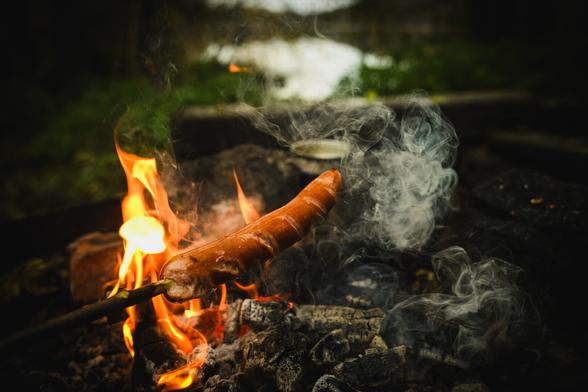 A photograph of a sausage being grilled over the embers of a camp fire. the sausage is held by a wooden stick, and the fire is mostly burned down to embers, but still burning a little.

In the out of focus background is some green grass and beyond that a smaller lake, surrounded by forest.