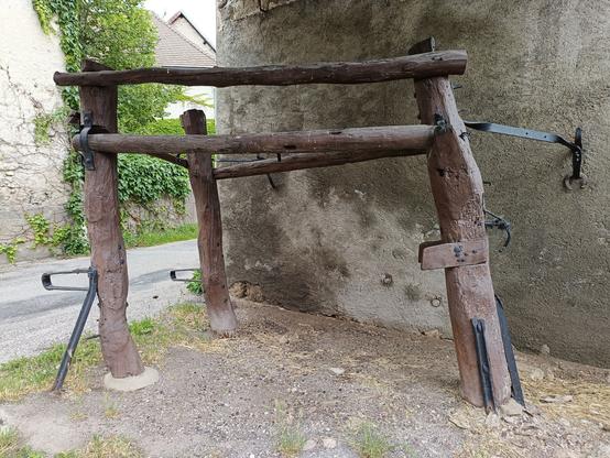 Sur ce modèle fixé à un mur, la structure comporte 3 montants en triangle fixé par des poutres en bois et des fers. Le sol est caillouteux et on aperçoit la rue derrière et d'autres maisons.