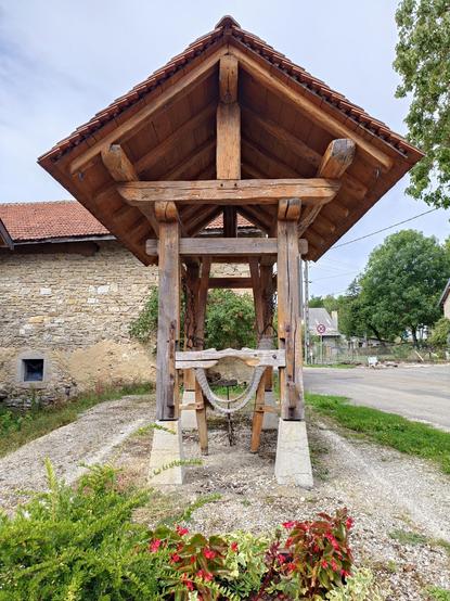 Ce modèle est composé de 4 montants en bois fixés sur des socles en béton, et il est surmonté d'un toit à 2 pans avec une charpente en bois et des tuiles rouges. La vue est prise dans le sens de la longueur, on voit des sangles et une structure en bois pour maintenir l'animal. 
Le travail est proche d'une rue dans un village dont on aperçoit des maisons.