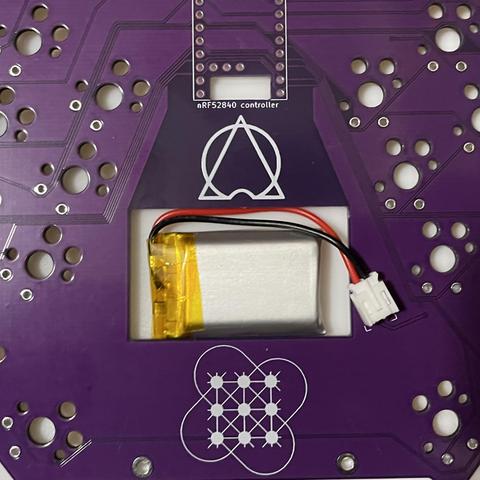 Photo of topside of Gamma Omega Hesse keyboard PCB (in purple) on the bottom case plate (white) with a small looking LP602030 LiPo battery in the cutout with plenty of room to spare connected to a JST connector which would be soldered in place later (and which impedes the battery compartment less than I feared). This is angled to match the case and nearest keys.