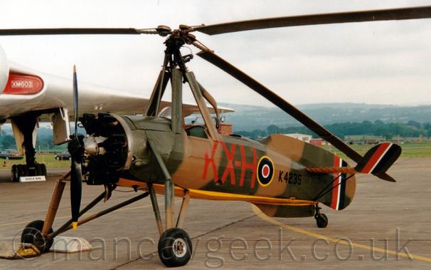 Side view of a World War 2-era autogyro parked facing to the left on a large, concrete apron.
The aircraft is covered in a brown and olive green camouflage pattern, with a yellow belly, with the large red letters "KX-H" below the two open, tandem cockpits, a blue, white, and red roundel outlined in yellow a bit further back, and the black serial"K4235" at the rear.
There are large red, white, and blue fin flashes on the tail, as well as on the up-turned tips of the horizontal stabiliser.
A large , twin-bladed rotary engine at the front is followed by long metal struts which hold the main landing gear, below more struts that hold a large, 3 bladed rotor overhead, just above the cockpit.
In the background, the white delta wing of a large, 4-engined jet bomber stretches halfway across the frame from the left, with views across the airfield and rolling, tree-covered hills in the distance on the right, under cloudy grey sky.