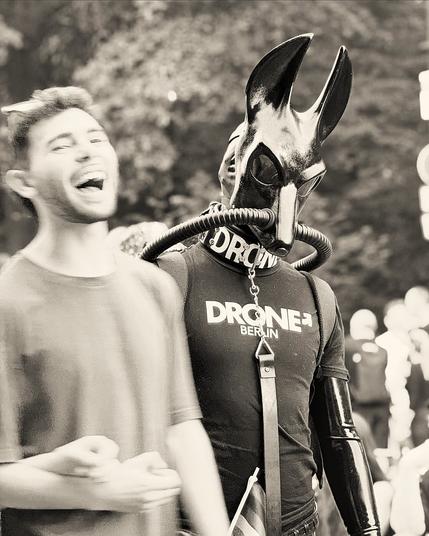 The jackal drone K-571 Codename "Obelisk" in its latex catsuit, jackal gasmask, drone shirt and with attached leash is in the center of the frame, hip upwards and in focus, giving the camera a view. Out of focus in the foreground, is a man cheering and laughing. In the background are more people from the CSD (gay parade) in Berlin 2025 visible as well as trees.
The drone is partly covered up by the man. It looks like a shadow of the man, the grim opposite of the happy person.
