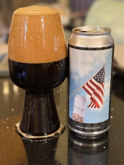A stout glass is overflowing (again) with dark beer and a huge dark tan head. The pint can is to the right showing an American flag flying on a blue sky background with an almond cookie adorned beer can pictured in the foreground