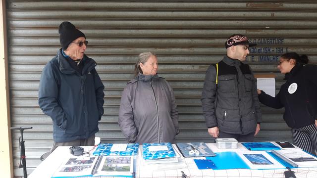 4 adultes à l'abri du crachin devant la devanture fermée du tabac ave la comm de l'asso posée sur une table haute pliante.