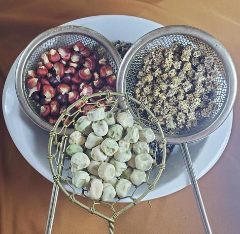 Three small sieves and a white plate displaying various dried seeds: red corn in one sieve, light brown seeds in another, and peas in a decorative wire basket sieve in front; all against a brown fabric background.
