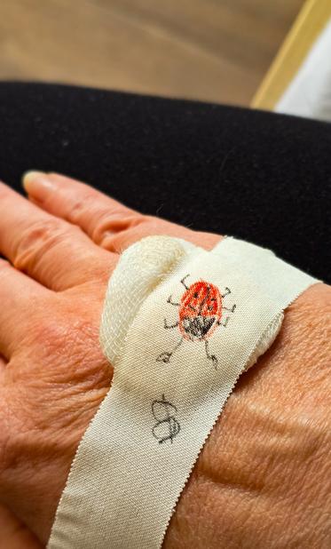 Eine Hand mit einem weißen Verband, auf dem ein kleiner roter Marienkäfer und eine fliegende Spur darunter aufgemalt sind. Der Verband klebt auf dem Handrücken, im Hintergrund ist dunkler Stoff zu sehen.