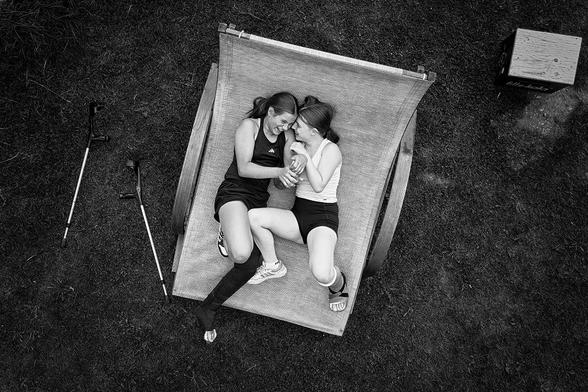 Zwei 12-jährige Mädchen mit Gipsen liegen nebeneinander auf einer großen Liege mit Stoffbespannung, aufgenommen aus der Vogelperspektive. Beide tragen sportliche Kleidung: ein Mädchen ein ärmelloses Oberteil und Shorts, das andere ein langärmeliges Oberteil und Shorts. Neben der Liege liegen zwei Gehhilfen auf dem Gras. Rechts oben im Bild steht eine kleine Holzkiste auf dem Rasen.