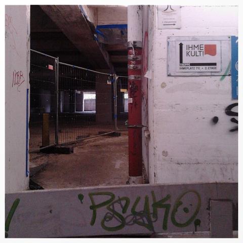 Foto aus dem Sockelgeschoss der Ruine des Ihmezentrums in Hannover-Linden. Vor einem improvisierten Durchgang, der einen Einblick in Dreck, Zerfall und endlose Stagnation der Scheinbaustelle gewährt, hängt ein Wegweiserschild für die Schönsprechagentur »Ihmekult«. Der Weg zu diesem Durchgang geht über eine im Bild nicht sichtbare Rampe, deren Geländer allerdings sichtbar ist. Das Geländer ist mit einem Graffito »Psyko« verziert.