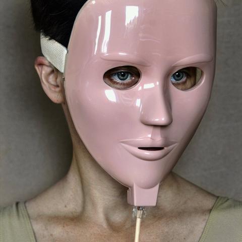 Una tipa indossa una maschera rosa probabilmente di vetroresina sulla quale si vedono dei riflessi. Le orbite della maschera sono  bucate e dunque si vedono gli occhi (azzurri) della persona che la indossa. La bocca della maschera è semi-aperta. La maschera è collegata alla testa, dietro, da un nastro bianco ben visibile sulla parte sinistra in alto della foto. Sotto il mento della maschera è collegato un cavo che sembrerebbe un cavo ethernet. La tipa è leggermente girata sul profilo destro e guarda fissa un punto sulla sinistra