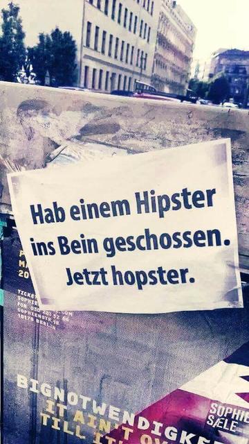 Das Bild zeigt ein an einer Wand aufgeklebtes, zerrissenes Papier. Es enthält Text in verschiedenen Schriftarten und Größen. Oben steht in großen, fetten Lettern "HAB EINEM HIPSTER". Darunter steht in kleinerer Schrift "INS BEIN GESCHOSSEN". Unten steht der Text "JETZT hopster". Der Hintergrund zeigt die Fassade eines Gebäudes mit dunklen Wänden und weißen Fensterrahmen. Es scheint sich um eine städtische Umgebung zu handeln.