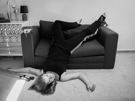 Ein 15-jähriges Mädchen liegt mit dem Rücken auf dem Boden vor einem dunkelgrauen Sofa. Ihr Oberkörper und Kopf ruhen auf dem Teppich, während ihre Beine auf dem Sofa ausgestreckt sind. Sie trägt ein schwarzes T-Shirt, schwarze Jogginghosen mit weißen Streifen an den Seiten und schwarze Socken mit weißer Schrift. Neben ihr auf dem Boden liegen zwei Krücken. Im Hintergrund steht ein kleiner Schrank mit einem Muster aus Kreisen und Rauten, darauf eine Vase mit einer Blume und eine Lampe.