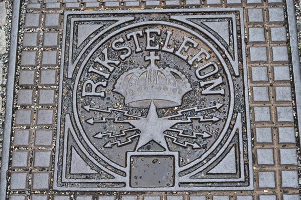 Gusseiserner Bodendeckel: in der Mitte ein fünfzackiger Stern, von dem nach links und rechts je vier Blitze ausgehen, darüber die schwedische Reichskrone, darüber im Halbkreis der Schriftzug 'Rikstelefon'.