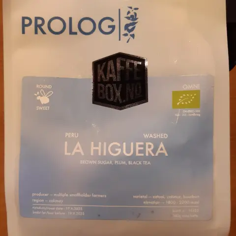The image shows a white coffee bag with several labels on it. At the top, there is a logo that reads "PROLOG." Below that, in large letters, it says "LA HIGUERA." The subtitle states "BROWN SUGAR, PLUM, BLACK TEA," indicating the flavor notes. There is additional information, including "PERU" and "WASHED," which likely refer to the origin and processing method of the coffee. The bag also has a label that reads "Kaffe Box No" and it includes details like "region: colasay," "varietal: catuai, catimor, bourbon," and "elevation: 1800 - 2200 masl."