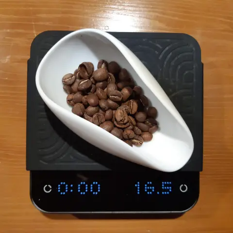 The image shows a digital scale on a wooden surface. On the scale, there is a white, curved bowl filled with brown coffee beans. The scale display shows two numbers: "00:00" on the left and "16.5" on the right, indicating a weight of 16.5 grams. The scale has a textured surface and is rectangular in shape.