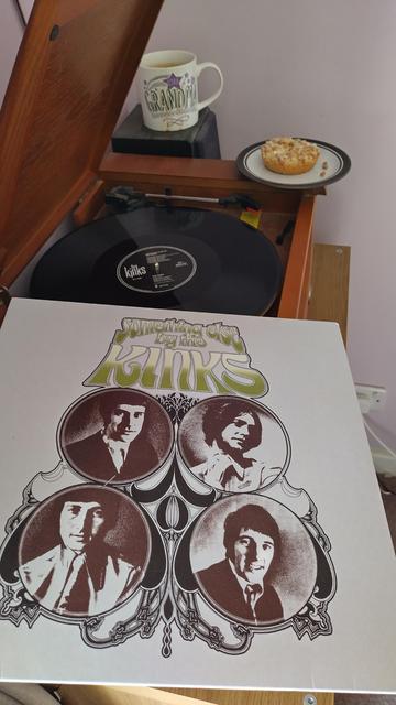 Record player with a 12" black vinyl record.
In the foreground is an album cover with four sepia photograph portraits of the band members.
Behind the player is a mug of coffee on a speaker.
On the corner of the player is a delicious mince pie.