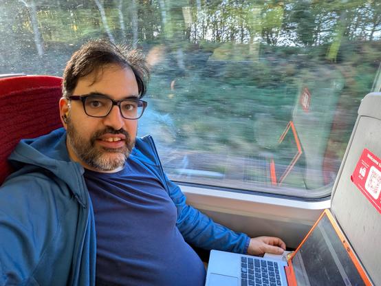 Keywan Selfie im Zug, am Fenster-Sitzplatz mit Laptop vor sich auf dem Klapptisch. Im Hintergrund vorbeirauschende Bäume