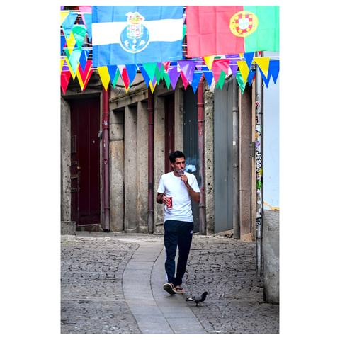 The image depicts a narrow, cobblestone street in an urban area. A man is walking down the street, wearing a white t-shirt, dark pants, and flip-flops. He is holding a can and is smoking a cigarette, blowing the smoke.
Above the street, colorful triangular pennant flags and larger flags are strung across, adding a festive atmosphere. The larger flags include the emblem of FC Porto, a Portuguese football club. The buildings lining the street have stone facades and closed doors, with some graffiti visible on the walls. A pigeon is seen on the ground near the man's feet. (draft by Mistral.ai, edited by author)