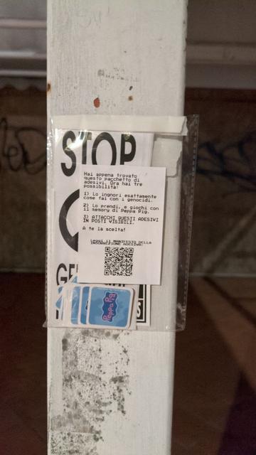 Pacchetto di adesivi su palo: 

“Hai appena trovato questo pacchetto di adesivi. Ora hai tre possibilità:

Lo ignori esattamente come fai con i genocidi.

Lo prendi, e giochi con il memory di Peppa Pig.

ATTACCHI QUESTI ADESIVI IN POSTI VISIBILI.

A te la scelta!

Leggi il MANIFESTO DELLA INSURREZIONE ADESIVA:”

Sotto il testo è presente un codice QR. Lo sfondo dell’etichetta è bianco.