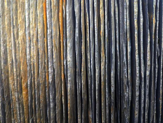 A close-up view of thin sheets of slate arranged vertically, showcasing their natural texture and layered appearance. The slate sheets display subtle variations in colour, ranging from dark grey and charcoal to hints of rusty orange and brown. The vertical alignment highlights the stone’s organic, striated patterns and uneven edges, creating a visually striking contrast between the darker and lighter tones. The lighting accentuates the depth and texture of the slate, emphasising its rugged, tactile quality.