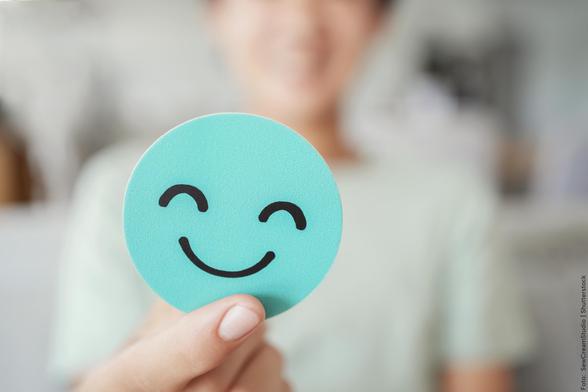 Foto: Nahaufnahme eines hellblauen, runden Smileys mit einer Hand im Vordergrund, unscharfe Person im Hintergrund.