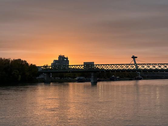 A sunset with the Danube river and the sky in a color gradient from orange to gray-blue and a bridge crossing the river in the middle of the image, behind it another bridge and some buildings to the left.