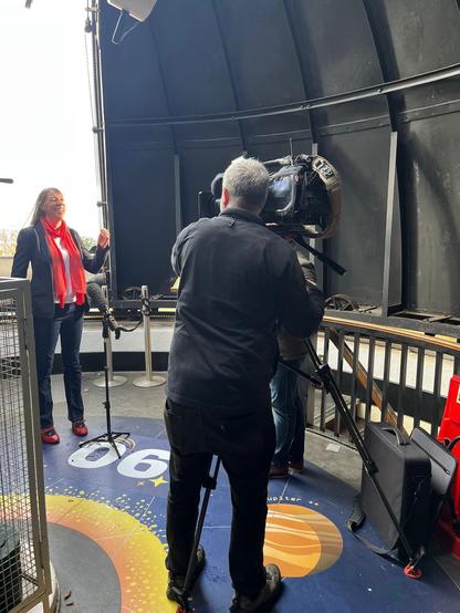 A film crew filming Catherine in a telescope dome.  She is in full flow, arms waving,  hair blowing, smiling/laughing, no doubt talking about how much she loves space.