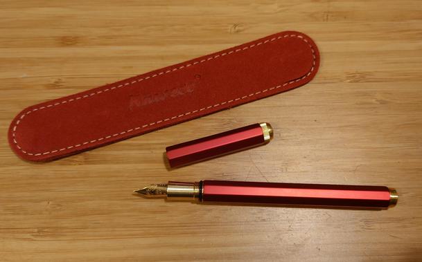 A metallic red fountain pain with an octagonal body and cap is lying on a desk, uncapped. The grip and nib, and finials on body and cap, are gold coloured. Next to the pen cap there's an oval red pen sleeve.