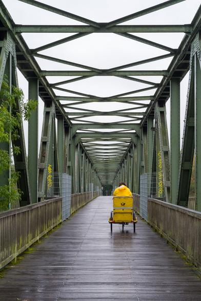 A mail carrier in yellow clothing rides a tricycle across a dark green bridge. The rider is only visible from behind, and the viewer's gaze follows his path across the bridge.
