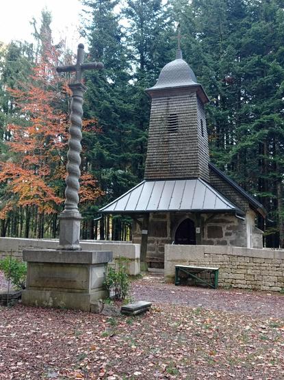 Ancienne croix de cimetière au fût torsadé, devant une chapelle au porche surmonté d'un clocher comtois bardé de bois.