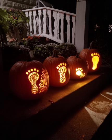 A row of carved pumpkins is displayed on a step at night, each illuminated from within. The designs include various footprints and patterns, creating a themed Halloween decoration. A white railing and flowers can be seen in the background.